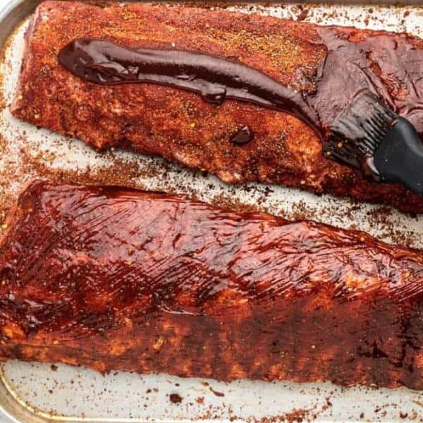 Two racks of ribs on a baking sheet—one plain, one brushed with barbecue sauce—a bowl of dry rub and a cup of sauce placed beside them.