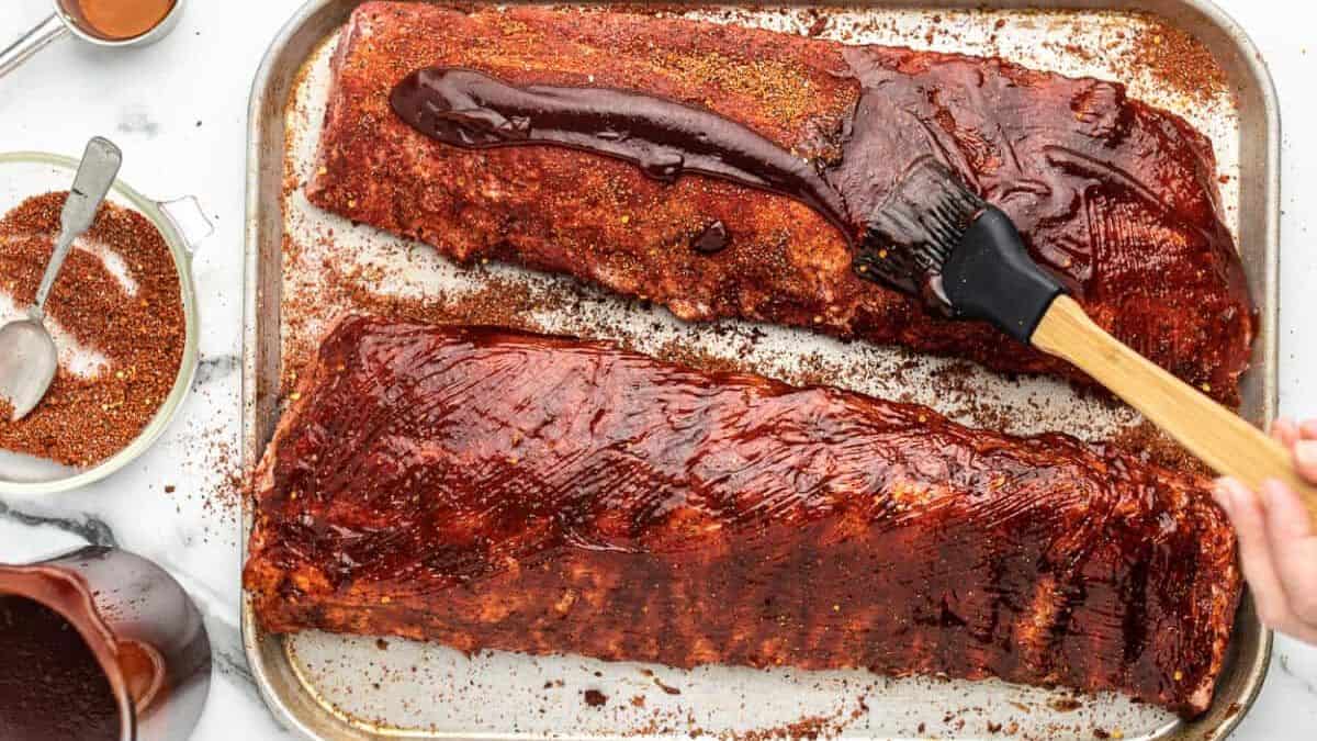 Two racks of ribs on a baking sheet—one plain, one brushed with barbecue sauce—a bowl of dry rub and a cup of sauce placed beside them.