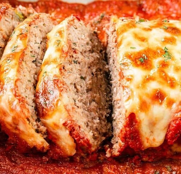 A close-up of sliced meatloaf topped with melted cheese and herbs, resting in a dish with tomato sauce.