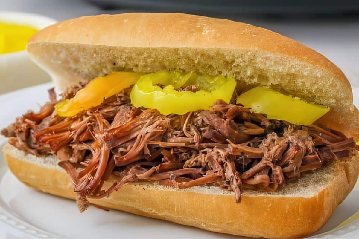 A sandwich roll filled with shredded beef and topped with sliced yellow peppers sits on a white plate.