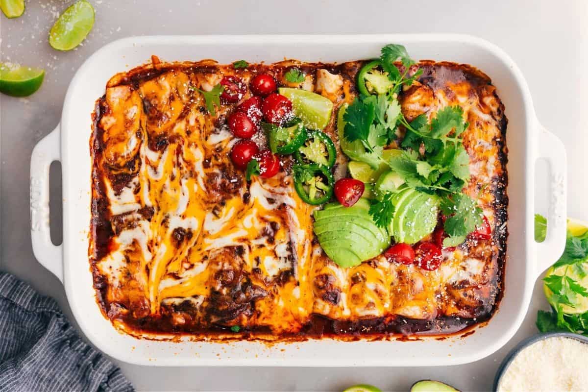 A casserole dish filled with cheesy baked enchiladas, topped with sliced avocado, lime wedges, jalapeños, cilantro, and cherry tomatoes. Some enchiladas are plain with melted cheese, while others have fresh toppings.