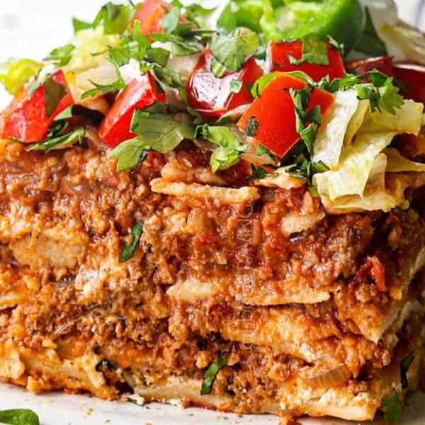 A close-up of a layered taco casserole topped with chopped lettuce, tomatoes, cilantro, and sliced jalapeño on a white plate.