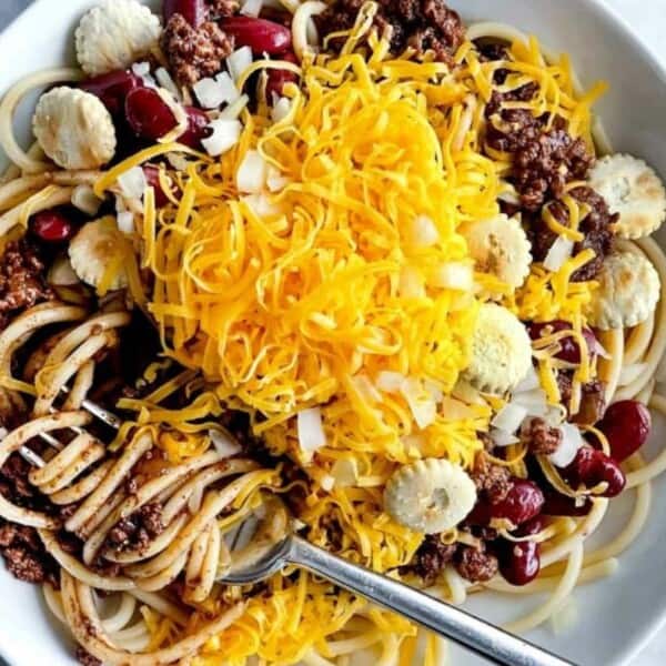 A bowl containing spaghetti, chili, kidney beans, shredded cheddar cheese, chopped onions, and oyster crackers; a fork rests inside the dish.