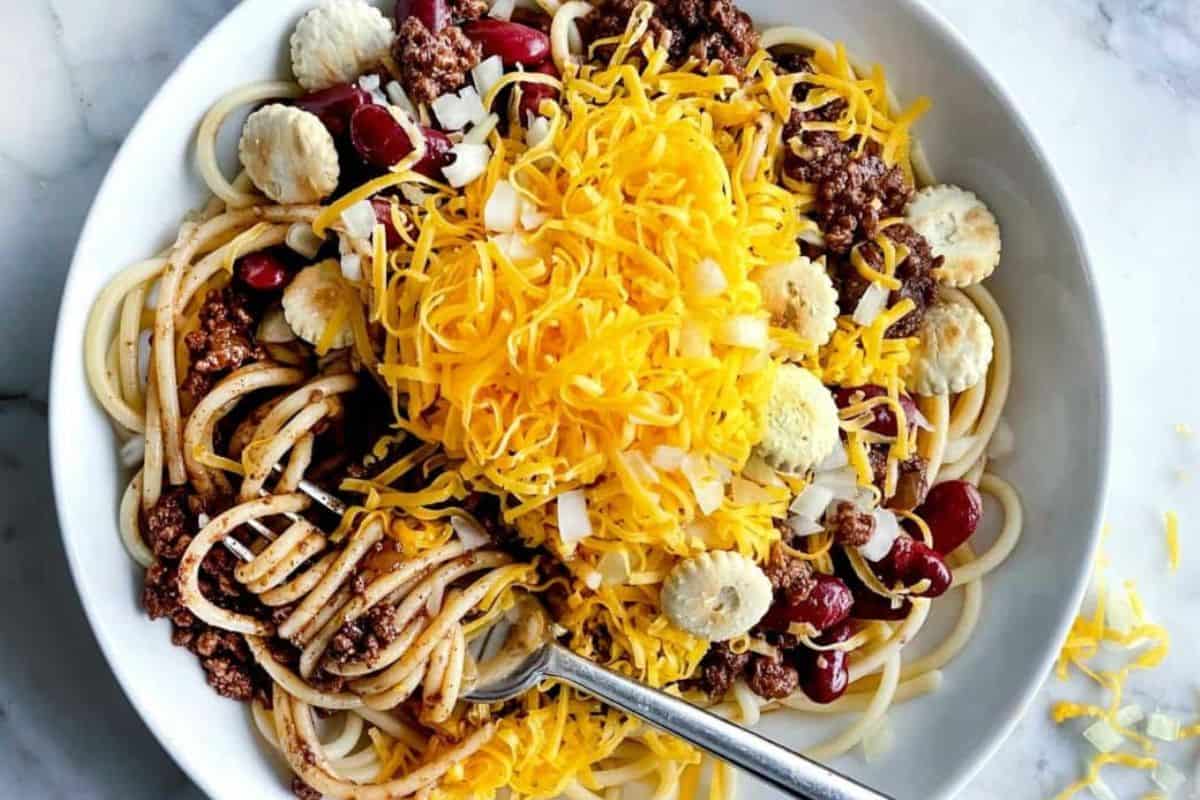 A bowl containing spaghetti, chili, kidney beans, shredded cheddar cheese, chopped onions, and oyster crackers; a fork rests inside the dish.