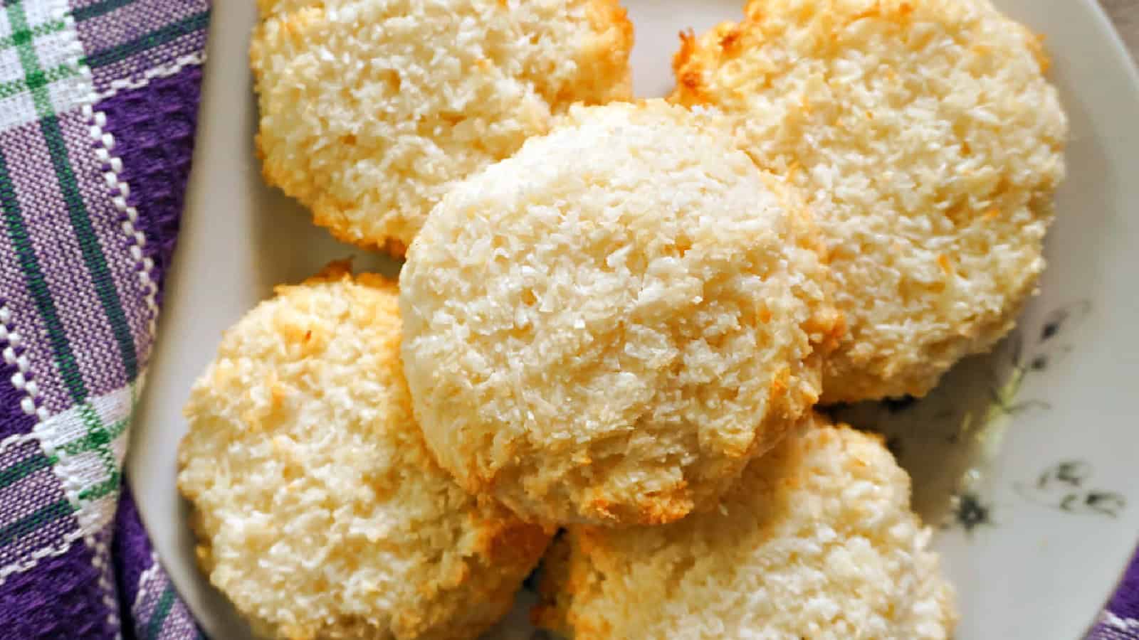 A plate of round, golden-brown coconut macaroons sits next to a purple and white checkered cloth.