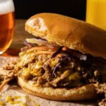 A pulled pork sandwich with melted cheese, pickled vegetables, and coleslaw on a bun is served on brown paper. In the background, there is a glass of beer and a yellow drink.