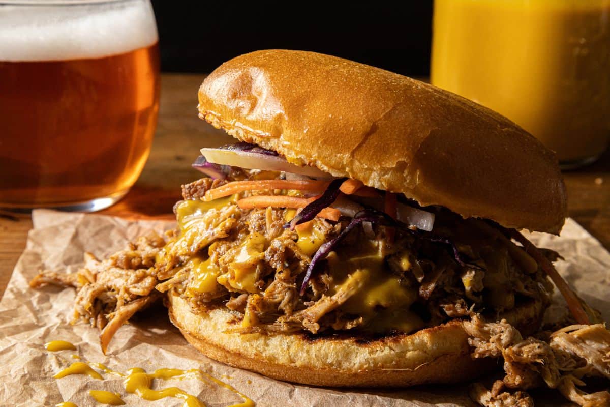 A pulled pork sandwich with melted cheese, pickled vegetables, and coleslaw on a bun is served on brown paper. In the background, there is a glass of beer and a yellow drink.