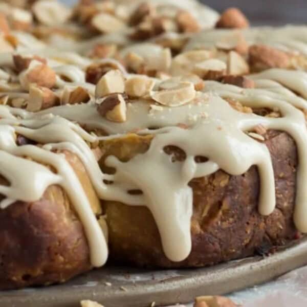 A round baked pastry with a glossy layer of white icing and sprinkled chopped nuts rests on parchment paper.