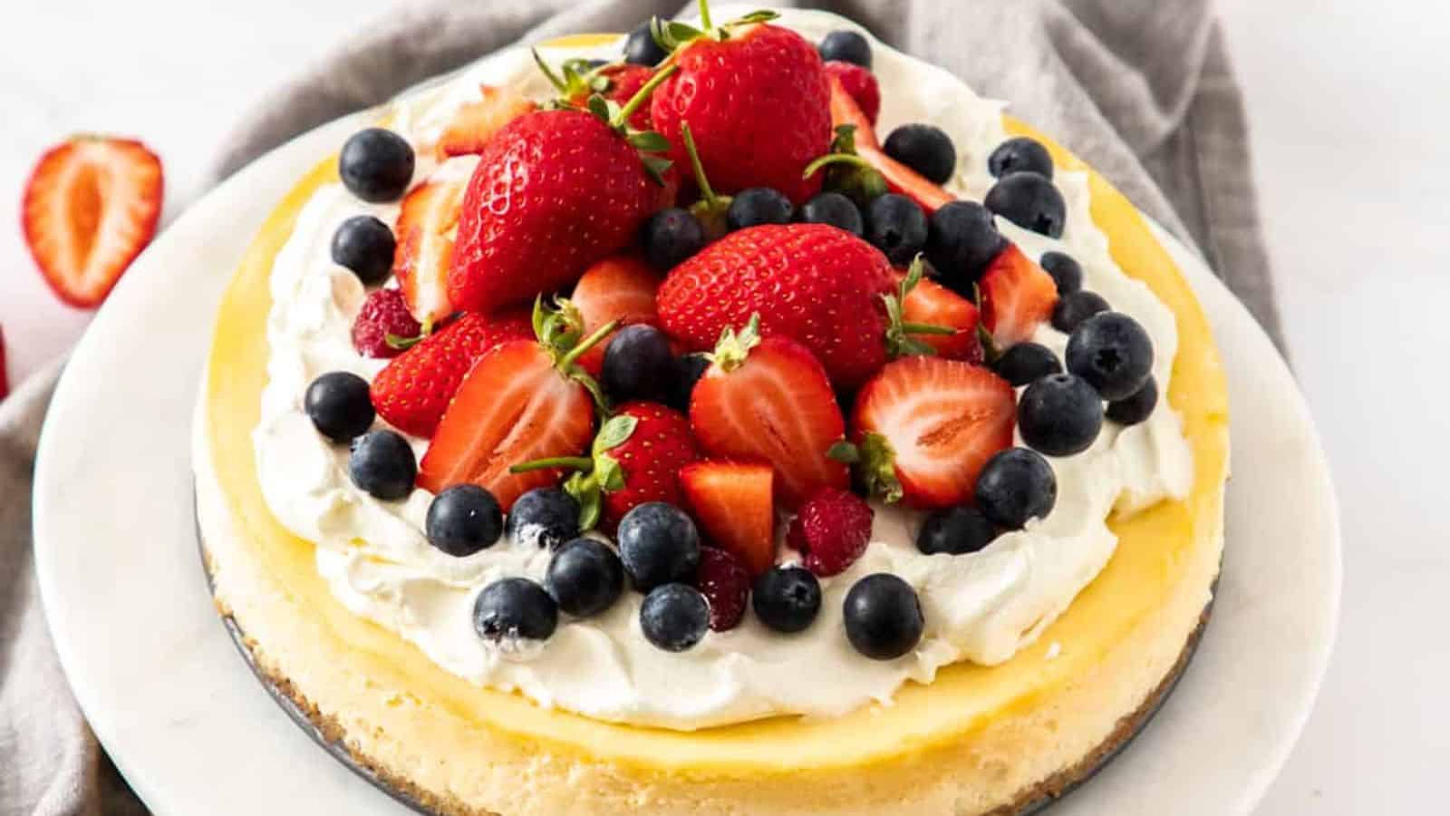 A slice of cheesecake on a white plate, topped with whipped cream, whole and sliced strawberries, blueberries, and raspberries.