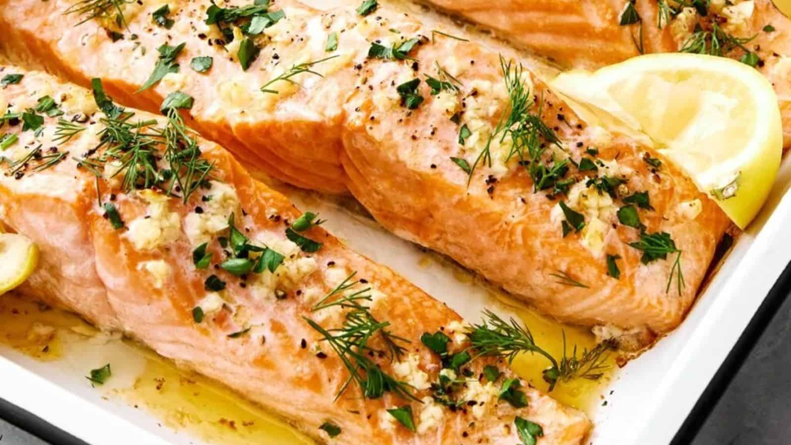 Three baked salmon fillets topped with minced garlic, black pepper, fresh herbs, and lemon slices on a white baking dish.
