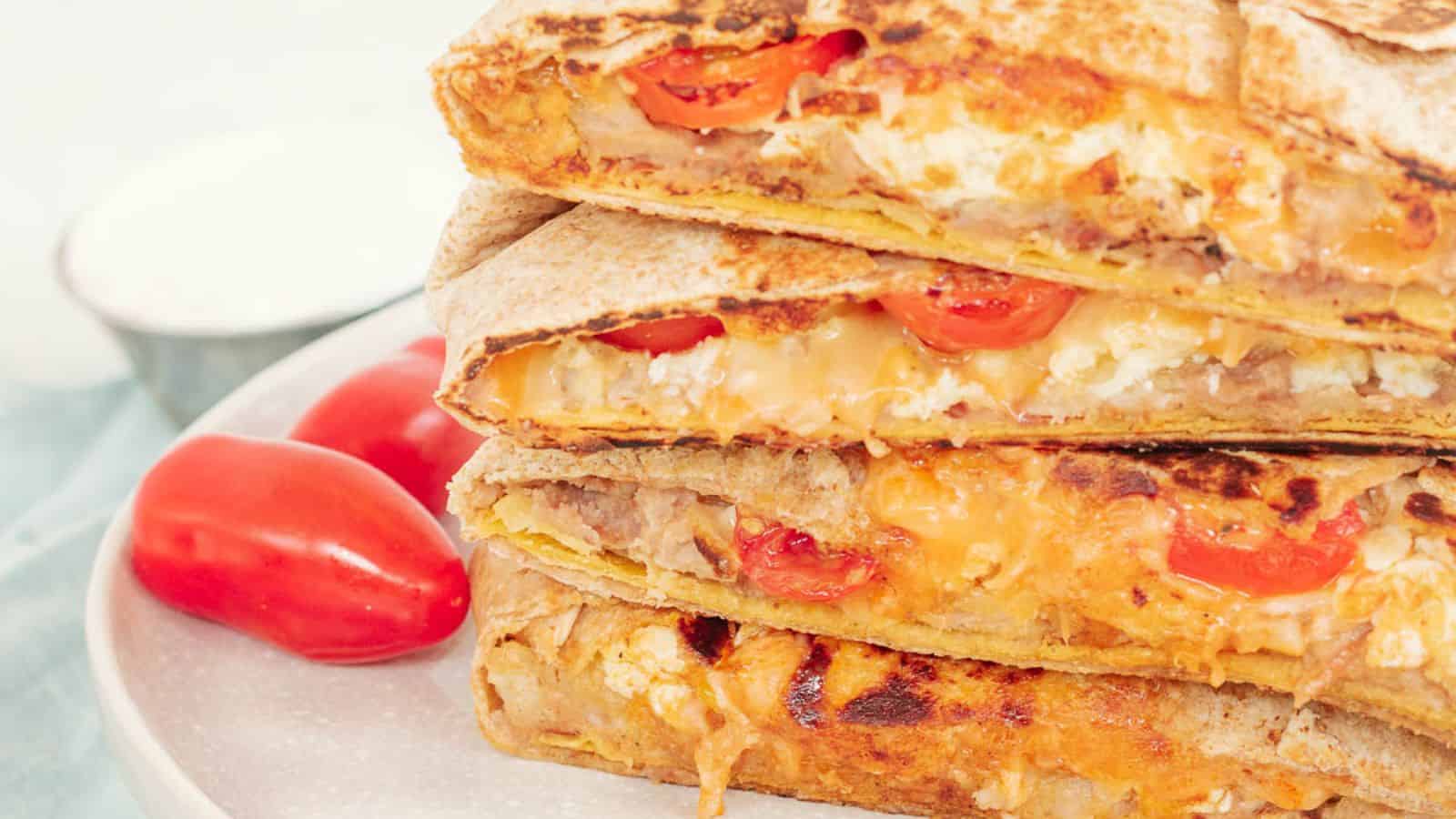 Three grilled quesadillas stacked on a plate, filled with melted cheese and diced tomatoes. Two grape tomatoes and a small bowl of sauce are in the background.