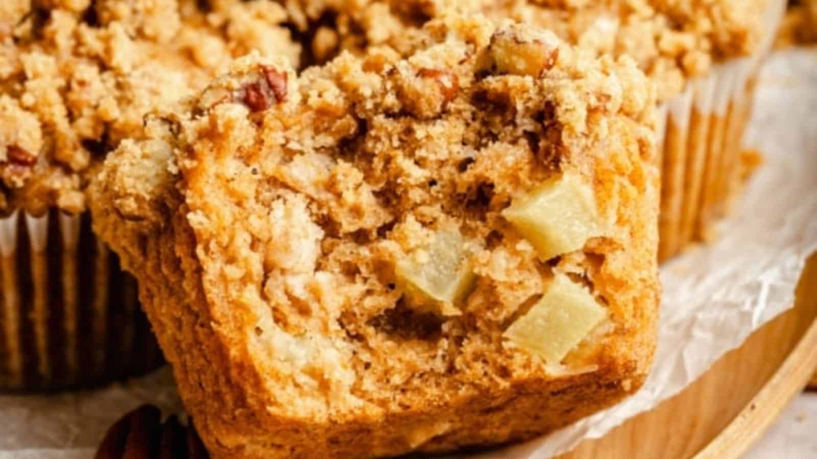 A muffin with a crumbly streusel topping and apple chunks sits on parchment paper, with another muffin blurred in the background.