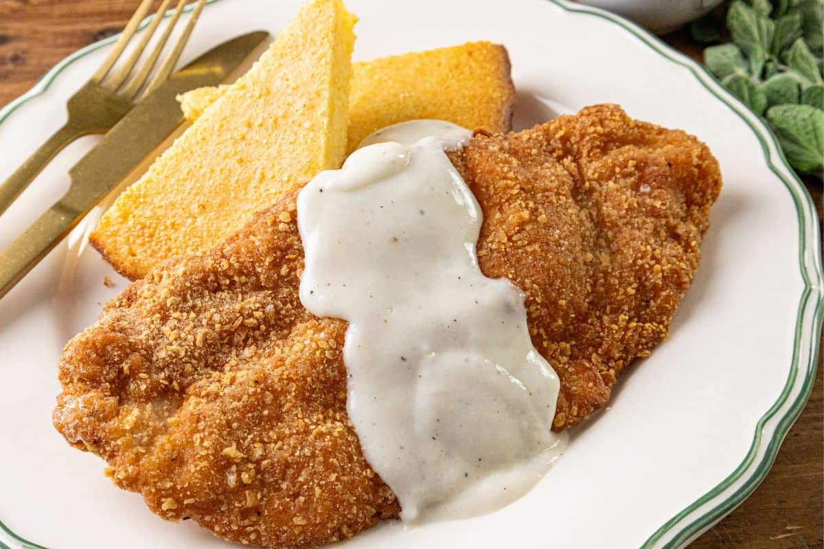 A plate with breaded fried steak topped with white gravy, two slices of cornbread, and gold-colored utensils.