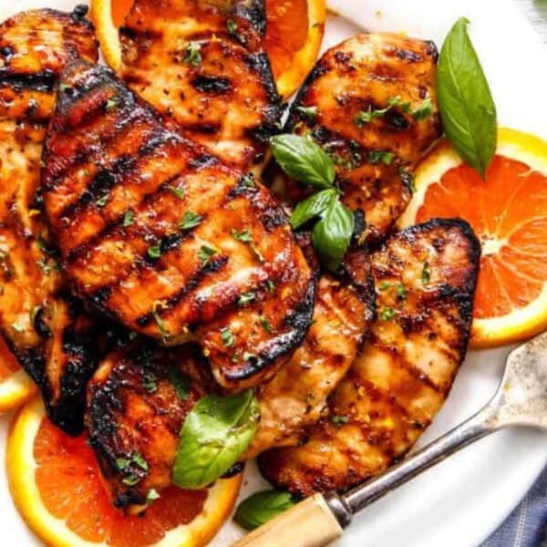 Grilled chicken breasts on a white plate, garnished with orange slices and fresh basil leaves, next to a fork.
