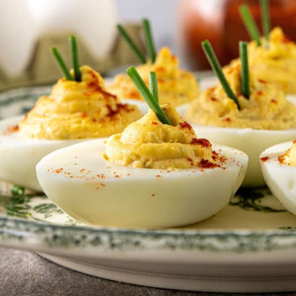 A decorative dish holds several halved deviled eggs with smooth, creamy yellow filling. The eggs are topped with small chopped green chives and a light dusting of red paprika. Some pieces of sweet pickle relish are visible on the filling.