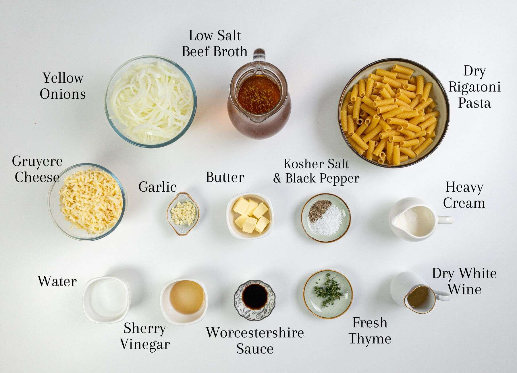 A top-down view displays dry rigatoni, sliced onions, blocks of cheese, butter, small bowls containing beef broth, heavy cream, garlic cloves, assorted spices, a sprig of thyme, vinegar, white wine in a glass, and water.