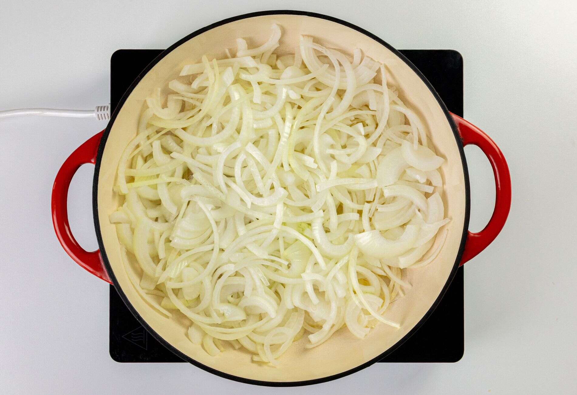 Thinly sliced onions in a large pot with red handles on a stovetop.