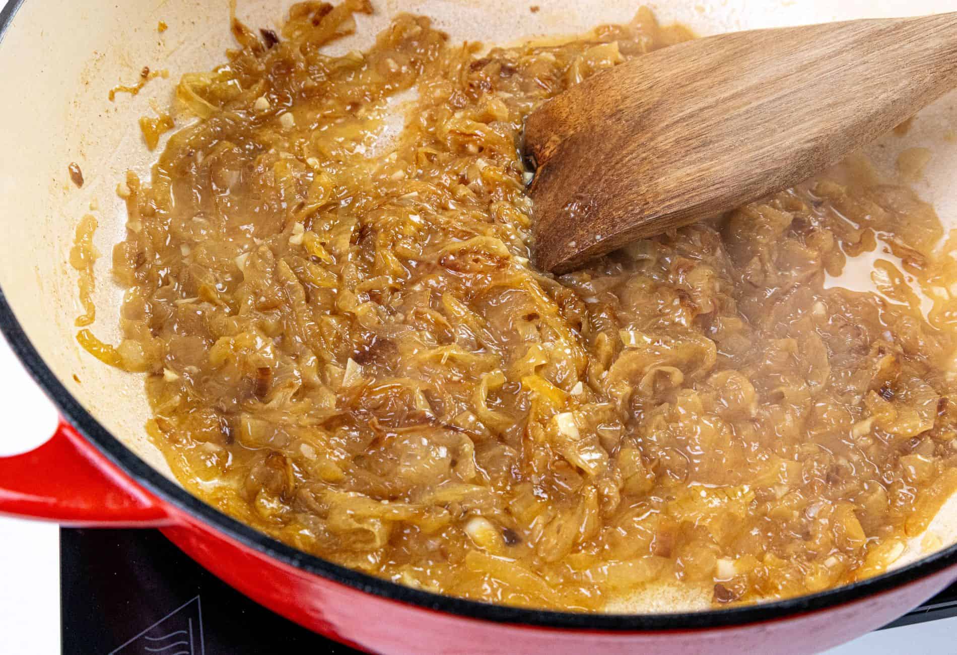 Caramelized onions in a red pot being stirred with a wooden spatula on a stovetop.