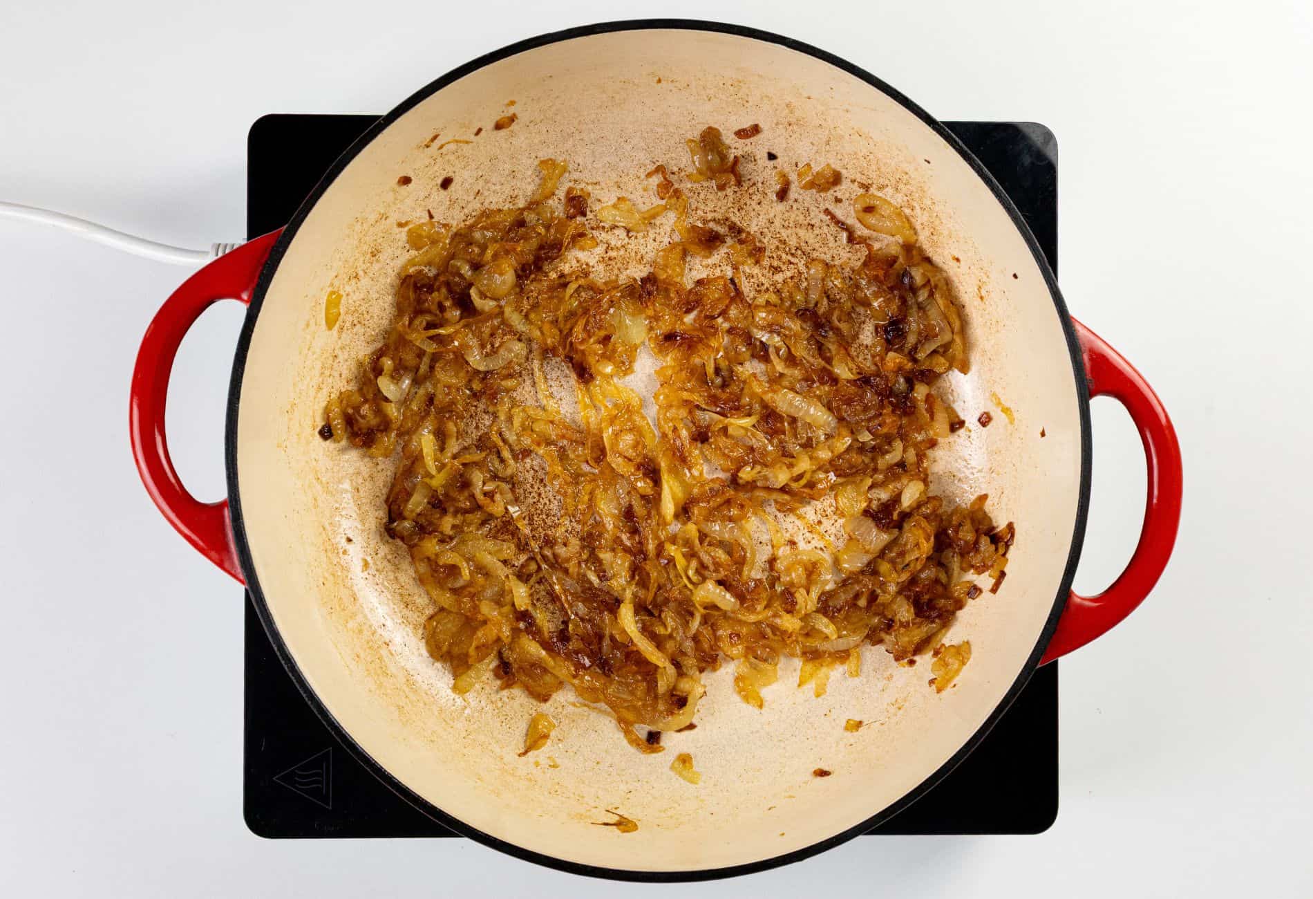 Sliced onions cooking in a red-handled pot on a black stovetop, with browned bits visible on the surface of the pan.