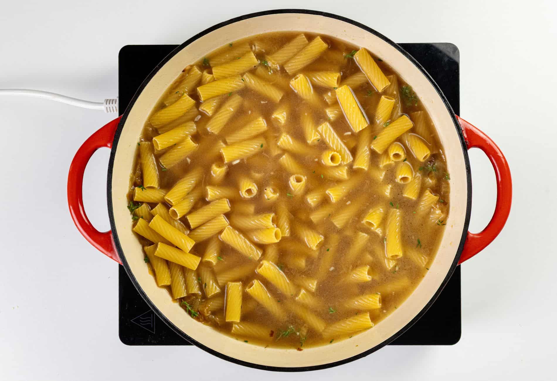 A large red pot on a stovetop, filled with uncooked rigatoni pasta, broth, and visible herbs.