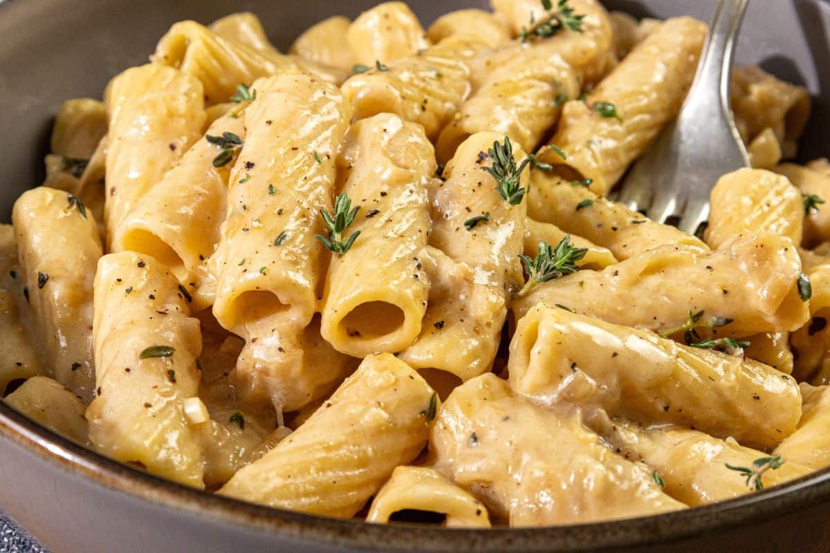 A bowl of rigatoni pasta coated in a creamy sauce, topped with black pepper and chopped fresh herbs, with a fork placed inside the dish.