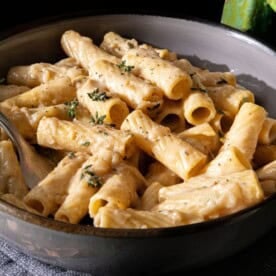 A bowl of rigatoni pasta coated in a creamy white sauce, topped with chopped fresh herbs, with a fork resting inside the bowl.