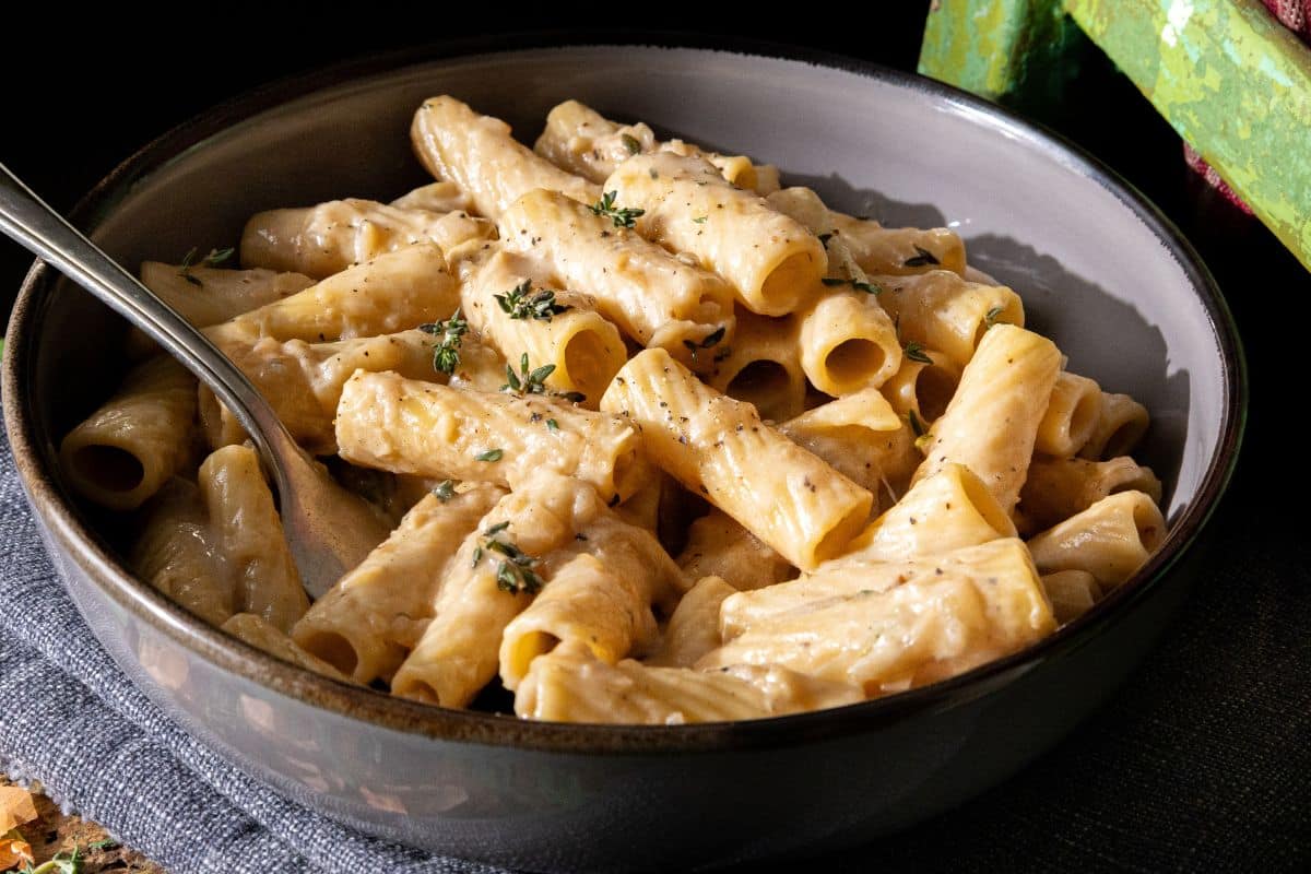 A bowl of rigatoni pasta coated in a creamy white sauce, topped with chopped fresh herbs, with a fork resting inside the bowl.