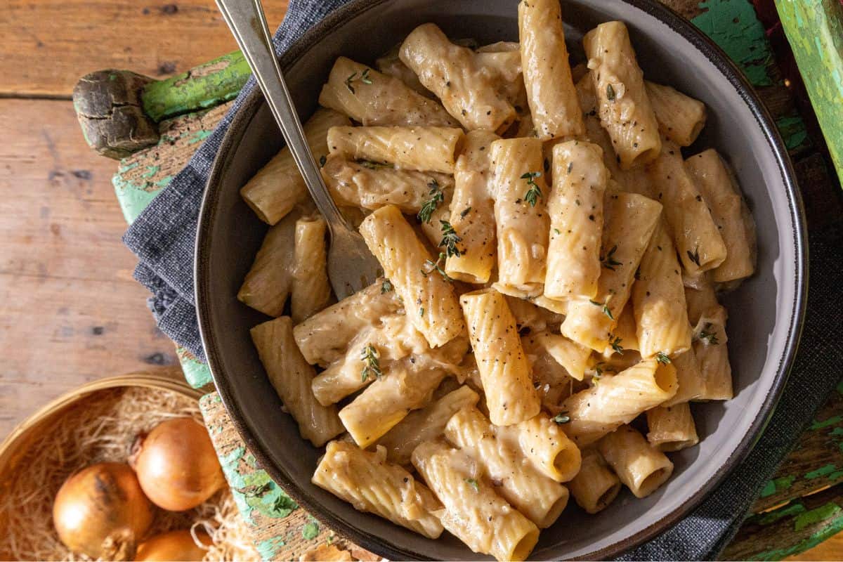 A bowl filled with rigatoni pasta in a creamy sauce is topped with fresh herbs and contains a fork. Beside the bowl, there are sliced onions and a small pile of grated cheese on a rustic wooden surface.