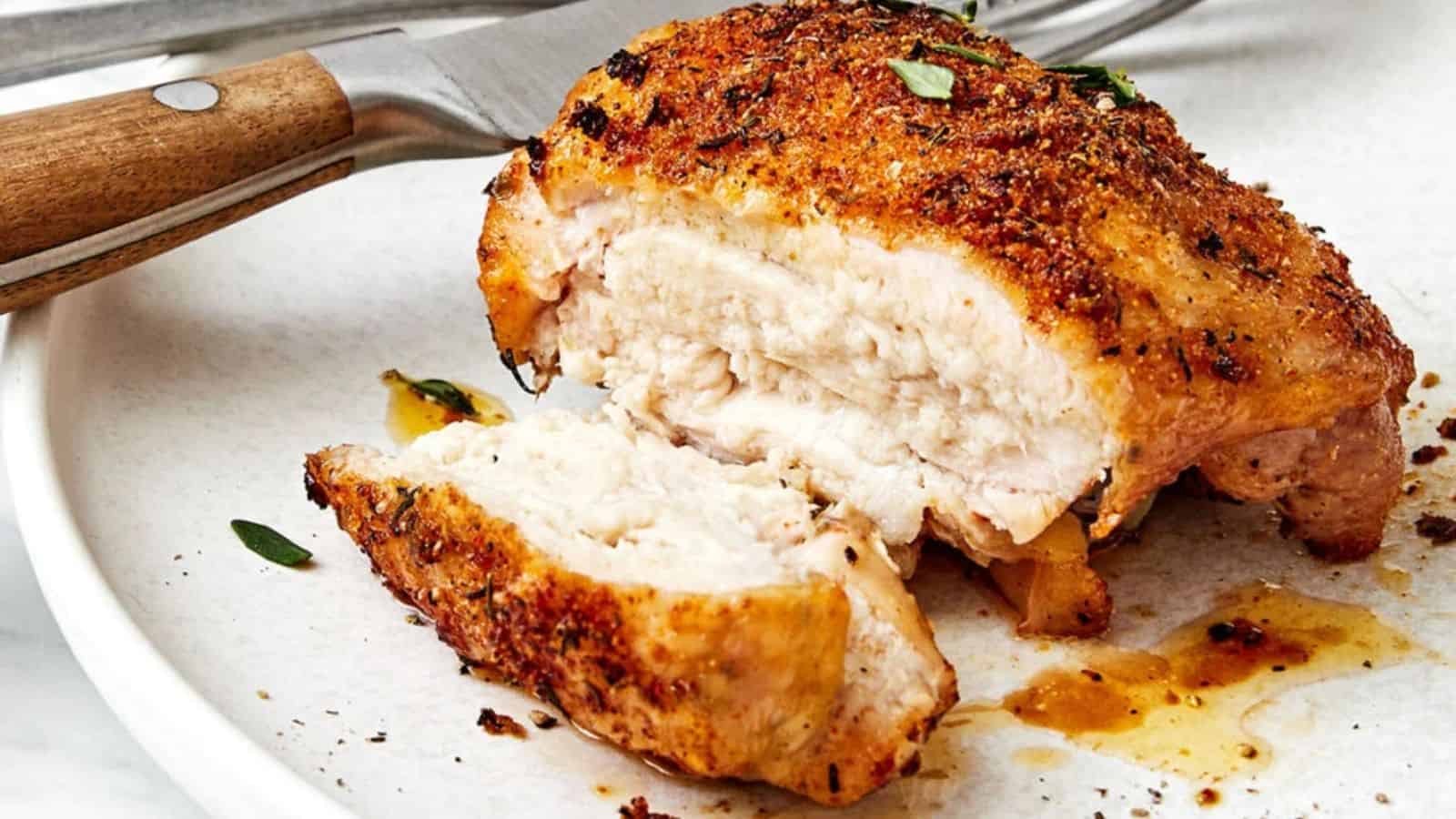 A cooked, seasoned chicken breast, partially sliced, sits on a white plate with a knife and fork placed nearby.