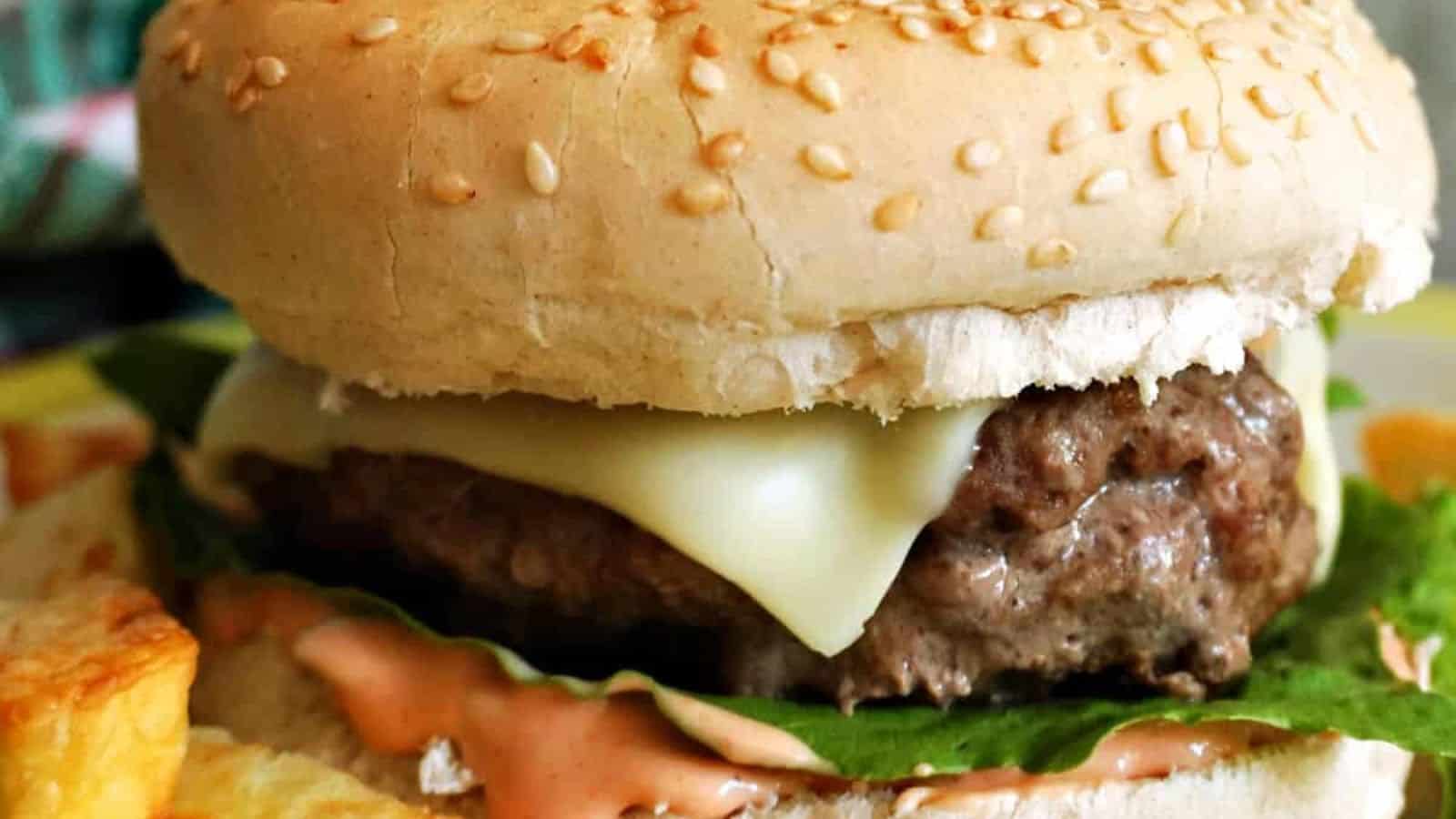 A sesame seed bun filled with melted cheese, lettuce, a burger patty, and sauce; a portion of fries appears to the side.