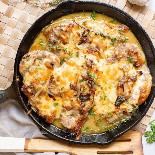 A cast iron skillet filled with baked chicken, melted cheese, caramelized onions, and herbs sits on a woven placemat.