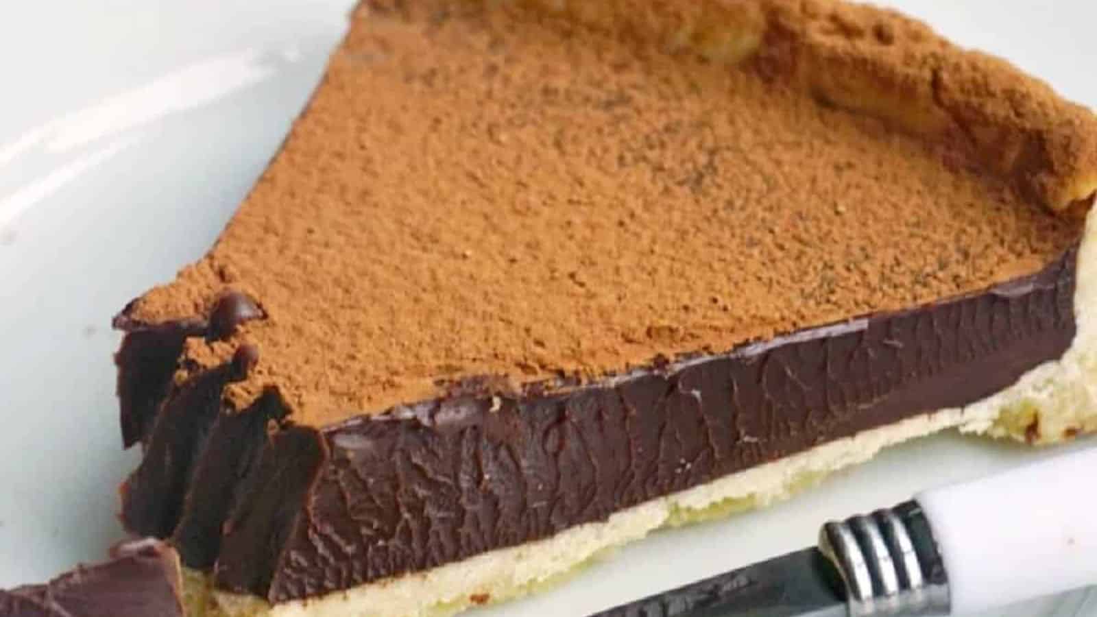 A slice of chocolate tart topped with cocoa powder, placed on a white plate alongside a knife.