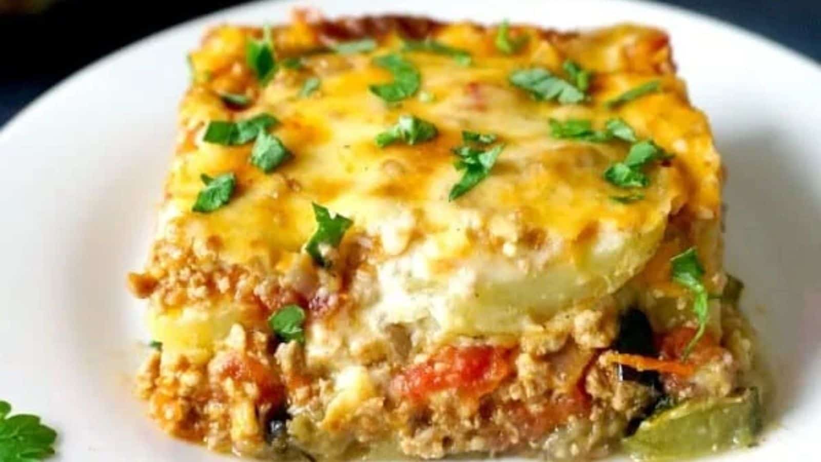 A slice of casserole featuring visible layers of ground meat, vegetables, and melted cheese, topped with chopped fresh herbs and placed on a white plate.