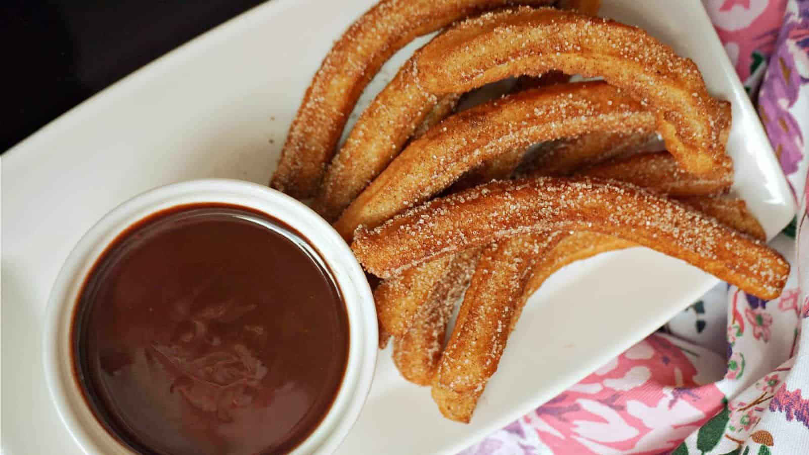 Five sugar-coated churros are arranged on a white plate beside a small bowl filled with chocolate sauce.