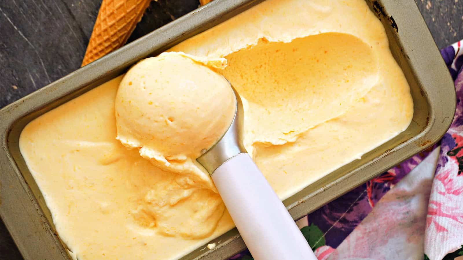 A metal ice cream scoop is in a container of creamy orange-colored ice cream. Beside it, there is a waffle cone and a floral-patterned cloth.