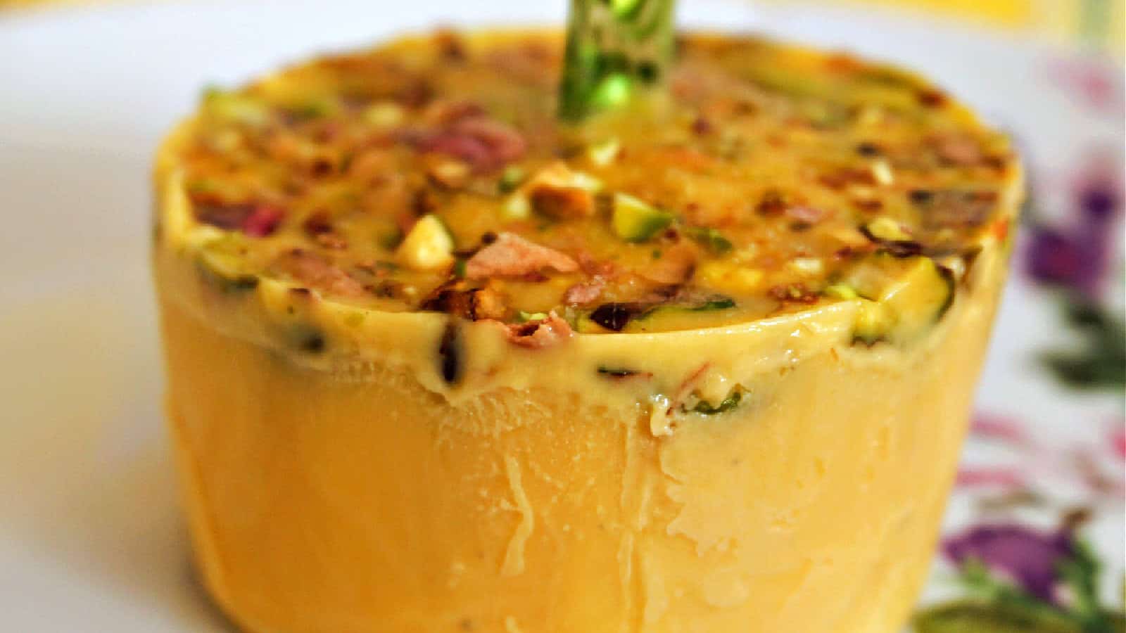 A round block of kulfi with a stick inserted in the center, topped with chopped nuts, placed on a white plate.