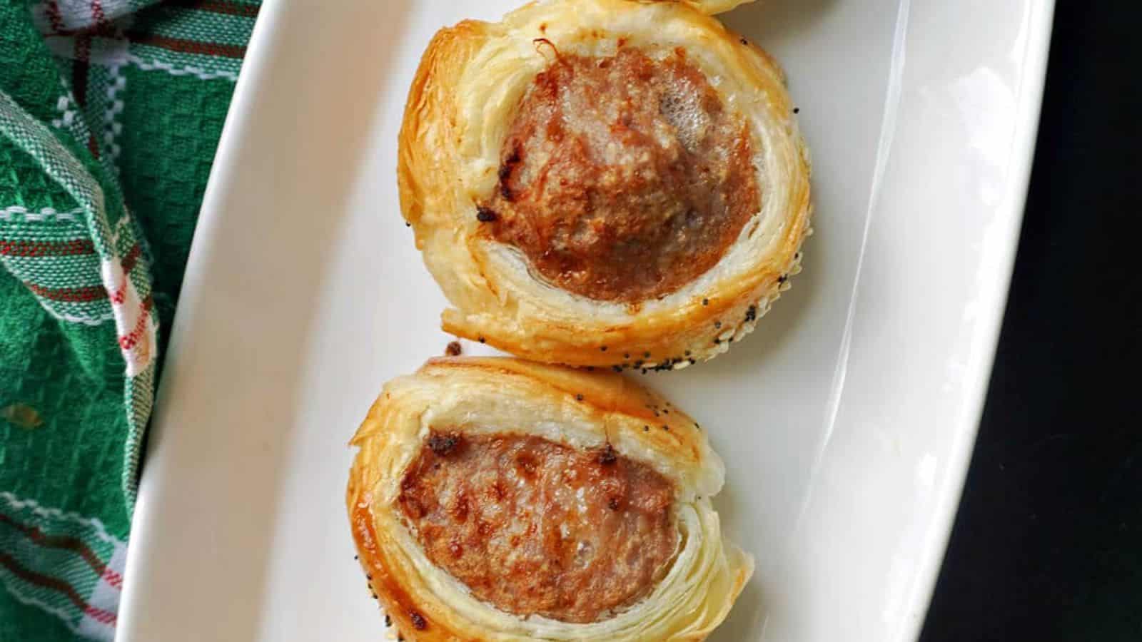 Two puff pastry-wrapped sausage rolls sit on a white rectangular plate beside a green and red cloth.