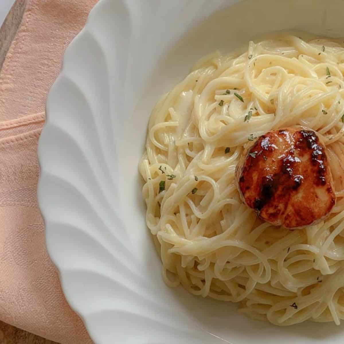 A white plate filled with rigatoni in a creamy sauce, topped with a grilled scallop and garnished with green herbs, rests on a wooden surface beside a peach-colored napkin.