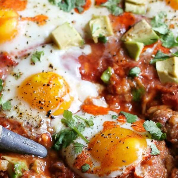 In the image, a skillet contains sunny-side-up eggs, chunks of avocado, tomato sauce, sprinkled herbs, and black pepper. A wooden-handled utensil rests on the side.