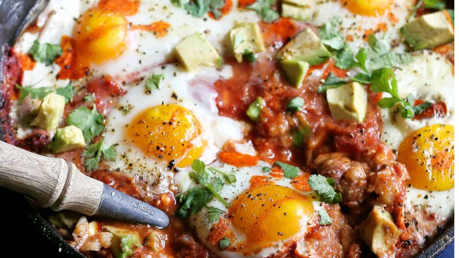 In the image, a skillet contains sunny-side-up eggs, chunks of avocado, tomato sauce, sprinkled herbs, and black pepper. A wooden-handled utensil rests on the side.