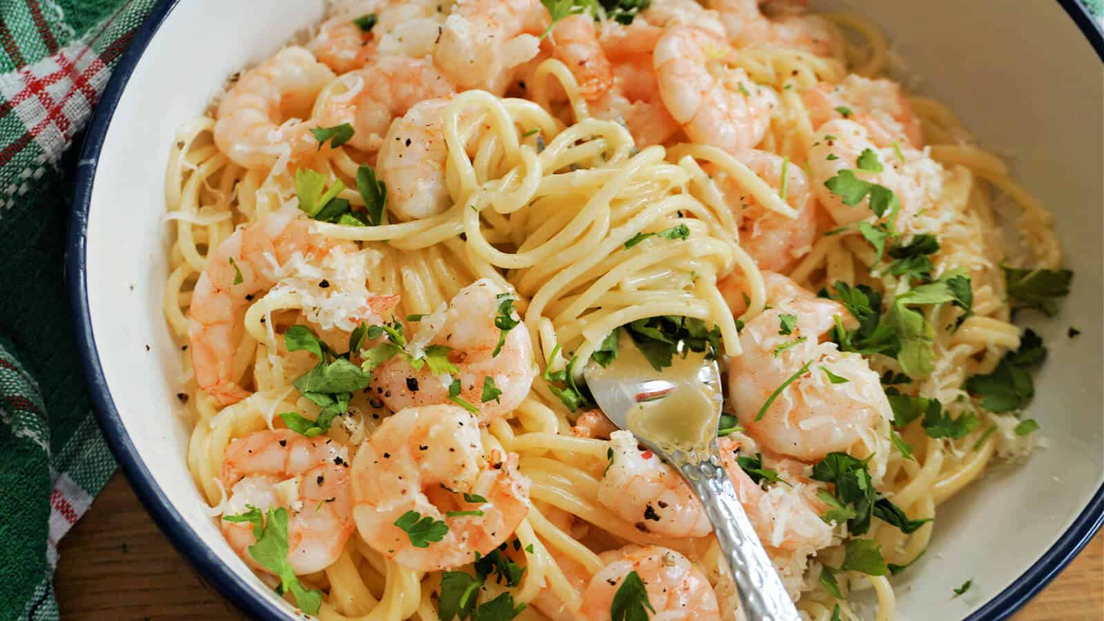 A bowl of spaghetti topped with cooked shrimp, grated cheese, chopped parsley, and black pepper, with a fork resting in the noodles.