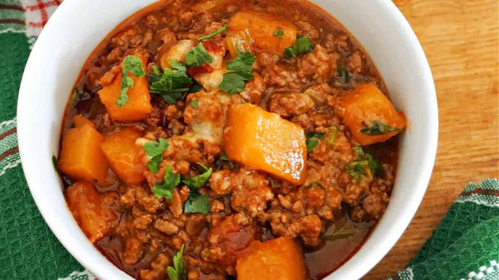 A white bowl filled with chili containing ground meat, tomato sauce, chunks of orange sweet potato, and chopped parsley on top.