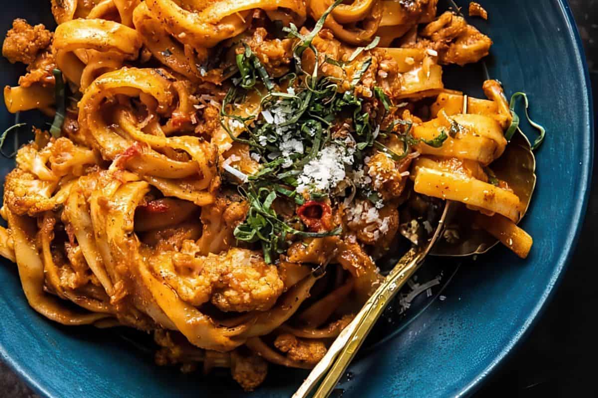 Tagliatelle pasta topped with tomato sauce, cauliflower florets, grated cheese, and fresh herbs in a blue bowl; a gold fork rests nearby.