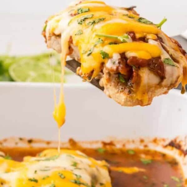 A spatula holds a cheesy chicken breast above a white casserole dish filled with melted cheese and sauce. Fresh herbs and a lime rest in the background.