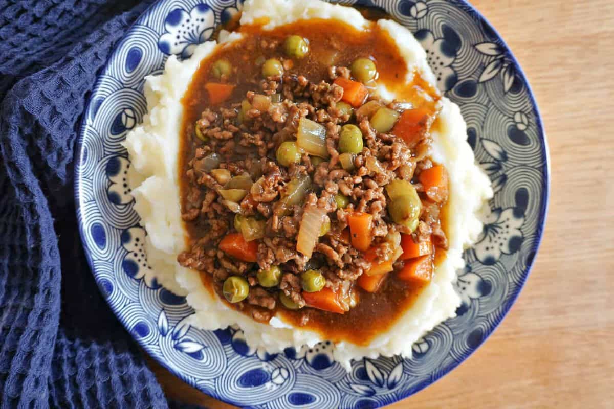 A blue patterned plate with mashed potatoes topped with ground beef, green peas, diced carrots, onions, and brown gravy.