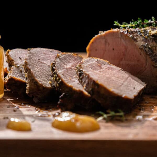 Slices of roast beef arranged on a wooden board with sprigs of herbs, next to a small jar containing a red sauce or jelly.