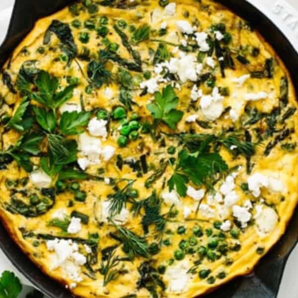 A black skillet containing a baked frittata topped with green peas, crumbled cheese, and fresh herbs sits on a white surface. Nearby are sprigs of herbs and a plate.
