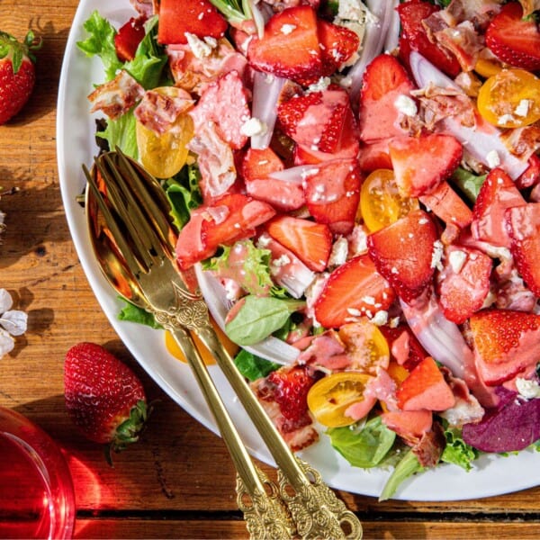 A salad with mixed greens, sliced strawberries, cherry tomatoes, bacon pieces, feta cheese, and pink dressing is presented on a plate with gold utensils on a wooden table.