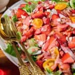 A plate of mixed greens topped with sliced strawberries, yellow cherry tomatoes, crumbled feta cheese, bacon pieces, and a pink dressing, with gold utensils placed beside the plate.