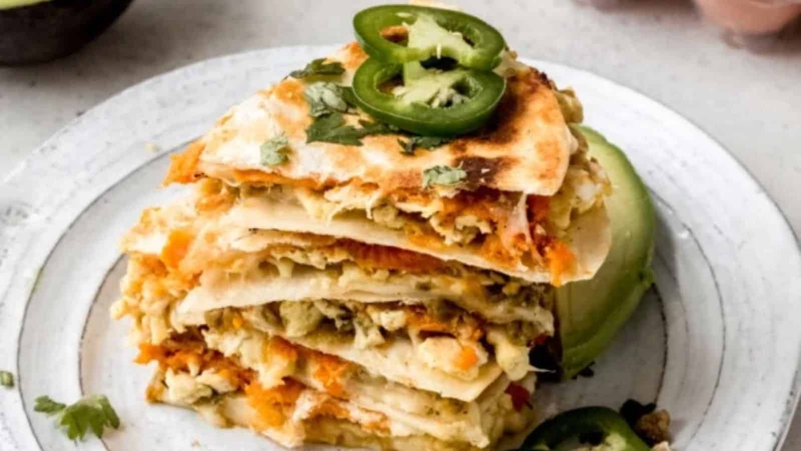 A stack of quesadilla slices with melted cheese and vegetables inside, topped with jalapeño slices and fresh cilantro, on a white plate.